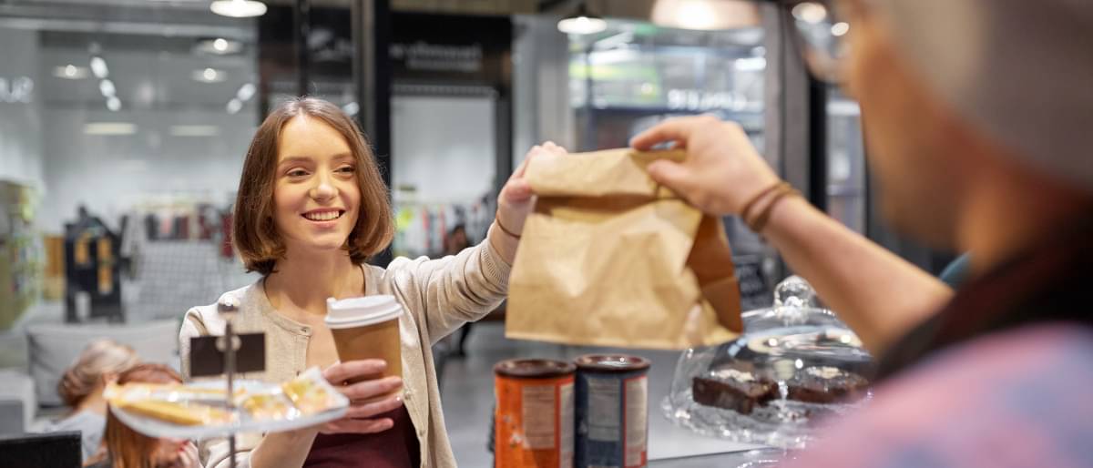 Eine junge Frau kauft in einem Coffee-Shop einen Becher Kaffee und erhält zusätzlich von der Verkäuferin eine Papiertüte mit Backwaren über die Verkaufstheke gereicht.