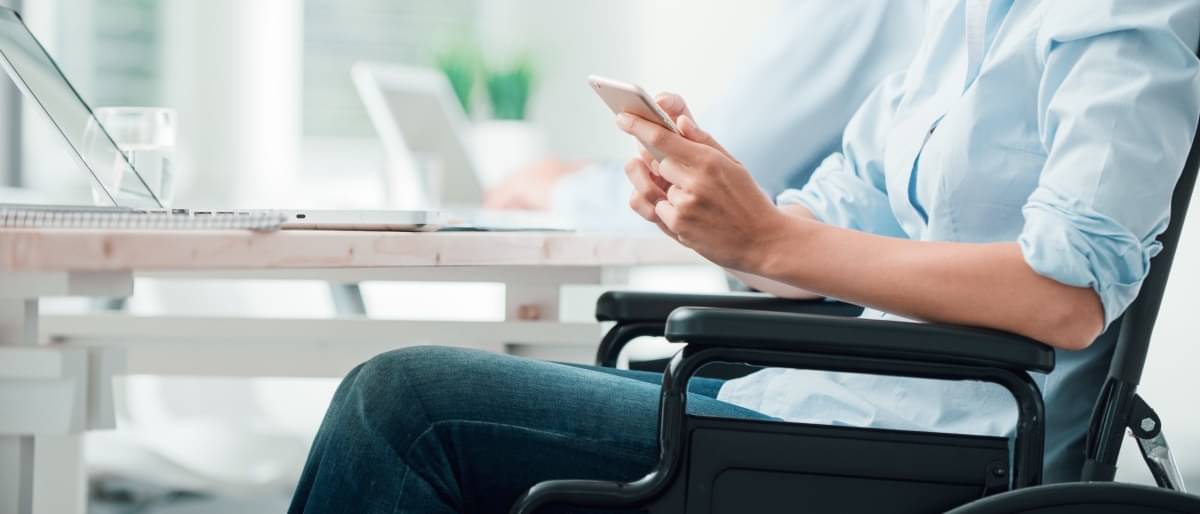 Frau sitzt an einem Schreibtisch im Rollstuhl und hat ein Tablet in der Hand 