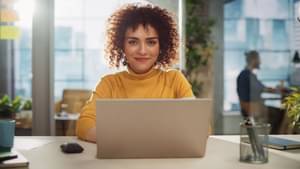 Eine Frau im gelben Rollkragenpulli sitzt vor ihrem aufgeklappten Laptop und lächelt in die Kamera.