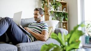 Mann mit Bart sitzt auf Sofa und ist über Laptop auf Sofatisch gebeugt.