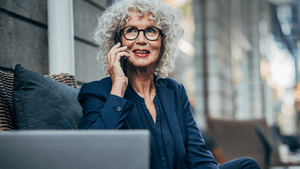 Mittelalte Frau mit grauen Haaren und Brille sitzt auf einem Sofa und träft einen blauen Blazer