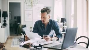 Junger Mann sitzt an Tisch vor Laptop und hat Unterlagen und Handy in den Händen.