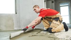 Ein Handwerker verteilt und glättet Estrich mit einer langen Holzleiste auf dem Boden eines Neubaus.