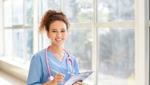 Eine junge Ärztin in blauem Arztkittel, mit einem Stethoskop um den Hals, hält ein Klemmbrett in der Hand und lächelt