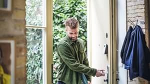 Junger Mann in grünem Cordhemd öffnet mit Blick nach unten die Wohnungstür und tritt ein. Laptop unter dem Arm.