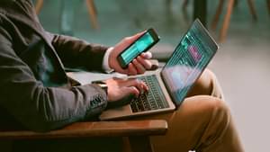 Ein Mann im Sakko sitzt auf einem Stuhl und studiert Diagramme auf seinem Smartphone und seinem Laptop.