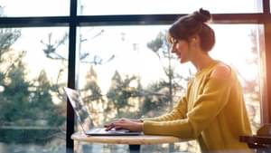 Frau sitzt vor dem Fenster mit Laptop. Die Sonne scheint ins Zimmer herein. 