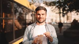 Junger Mann mit Coffe to go Becher steht vor einem Bus und lächelt in die Kamera.