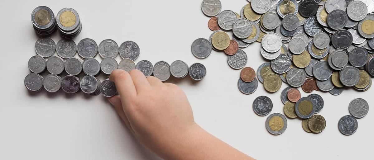 Viele Münzen liegen gehäuft auf einem weißen Tisch. Eine kleine Hand nimmt die Münzen von rechts und stapelt sie auf der linken Seite.