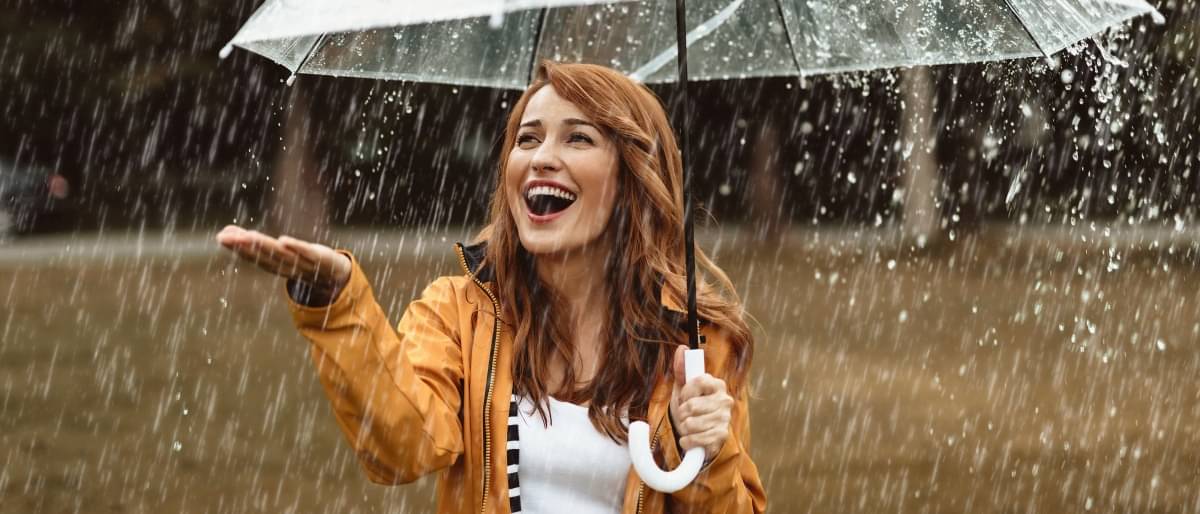 Junge Frau steht lachend mit transparentem Schirm im Regen und streckt ihre Hand aus