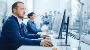 Eine Frau und ein Mann sitzen konzentriert vor ihren PCs und schauen auf ihre Bildschirme, im Hintergrund steht eine weitere Person und liest in einer Unterlage.