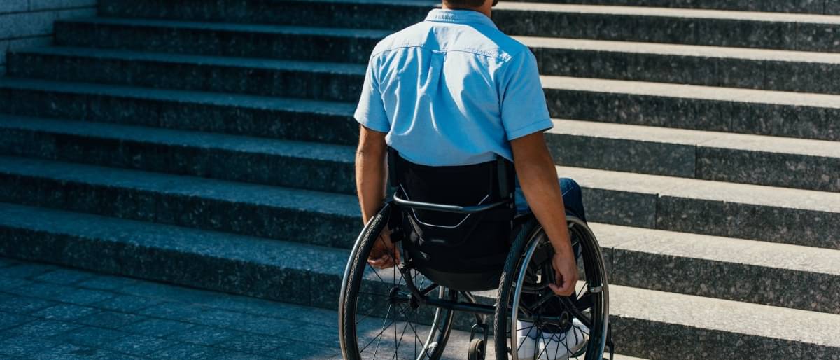 Ein junger Mann im blauen Hemd steht in seinem Rollstuhl vor einer Steintreppe