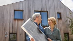 Eine Frau und ein Mann stehen vor einem Haus mit Holzvertäfelung, lächeln sich an, während der Mann eine Solarpanel in den Händen hält.
