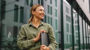 Eine Unternehmerin läuft mit ihrem Laptop im Arm an einem modernen Bürogebäude vorbei.