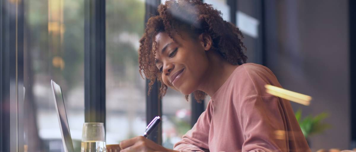 Eine junge Frau sitzt lächelnd vor ihrem Laptop und macht sich Notizen mit einem Kuli.