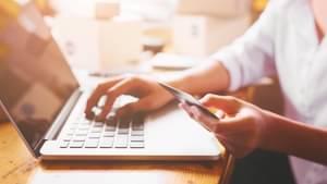 Eine Frau sitzt am Tisch und tätigt einen Online Einkauf an ihrem Laptop. In der linken Hand hält sie eine Kreditkarte.