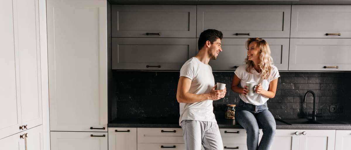 Ein junges Paar sitzt lachend in Ihrer neuen modernen Küche auf der Küchenzeile. Eine Immobilie lieber jetzt oder später kaufen? Es lohnt sich, die Baufinanzierung trotz Zinswende noch heute zu sichern.