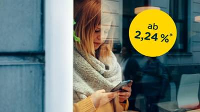 Junge Frau steht hinter bodentiefen Fenster mit Schal und gelbem Oberteil. Sie hält Handy in der Hand und tippt auf dem Bildschirm: Onlinekredit ab 2,24% p.a.*