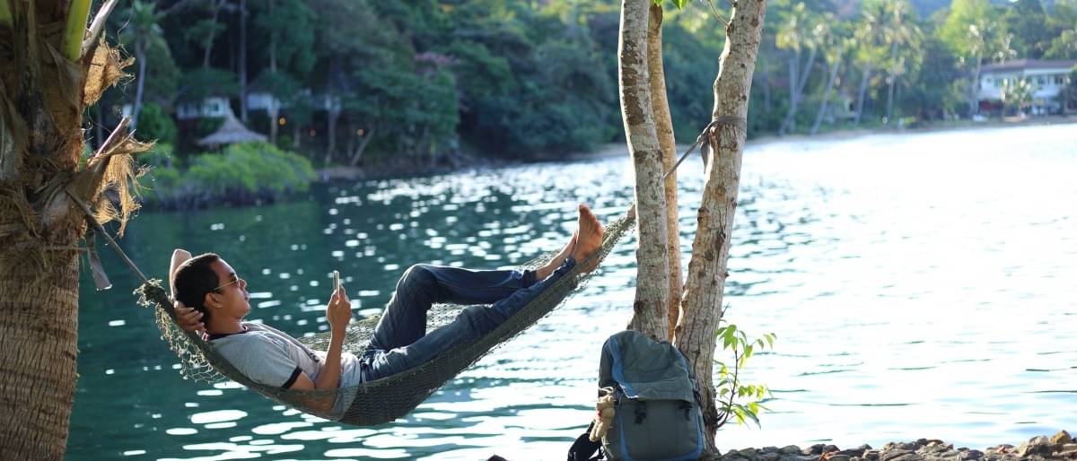 Ein Mann liegt entspannt in einer Hängematte, die zwischen zwei Palmen aufgehangen wurde, da er dank passiven Investments ausgesorgt hat. Im Hintergrund sieht man einen ruhigen See und einen Tropenwald.