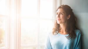 Junge Frau mit lockigem Haar, die ein Tablet in der Hand hält und aus dem Fenster in die ferne schaut.