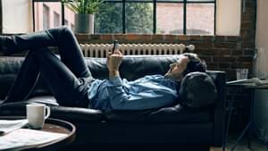 Eine dunkelhaariger Mann liegt auf dem Sofa und schaut lächelnd auf sein Handy