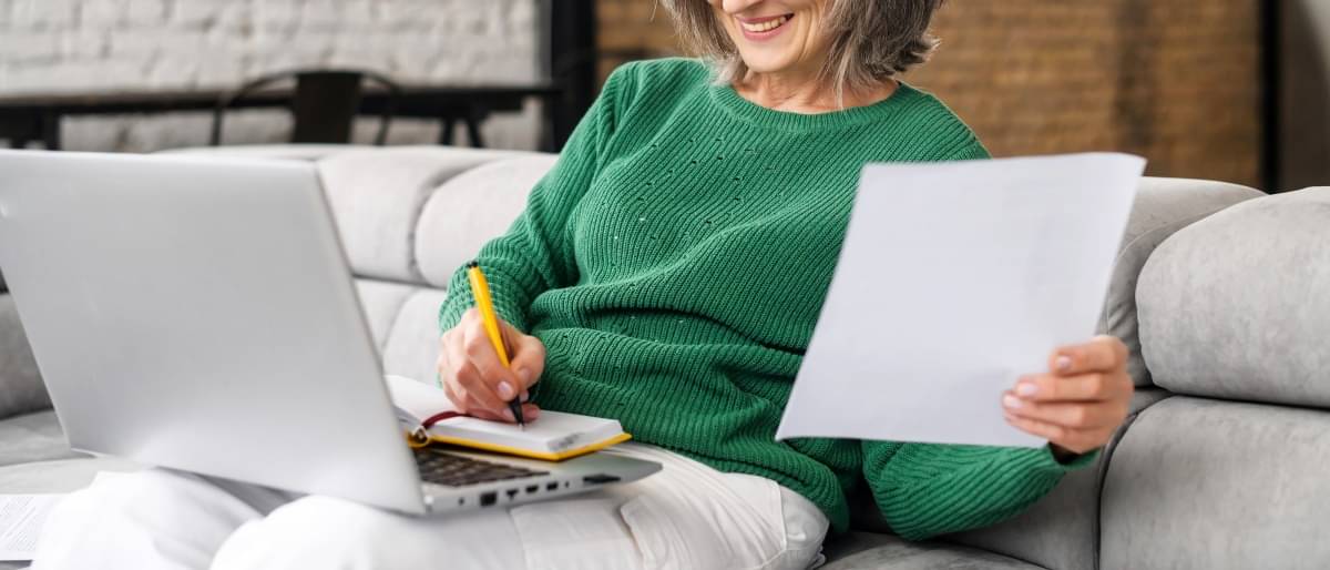 Eine ältere Frau mit grauen Haaren sitzt lächelnd mit Laptop auf dem Sofa und macht sich Notizen