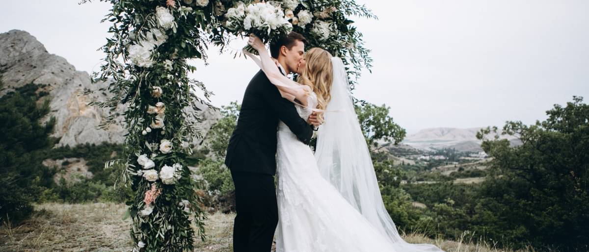Eine junge Braut im weißen Kleid und mit langem Schleier und ihr junger Ehemann stehen auf einem Hügel unter einem Rosenbogen und küssen sich. Dabei hält die Braut ihren Brautstauß in die Luft.