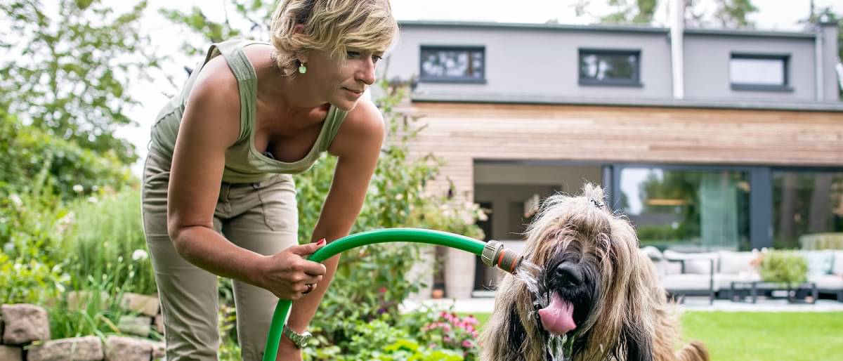 Frau mit kurzen blonden Haaren steht in Garten vor einem Haus und spritzt Hund neben ihr mit den Gartenschlauch ab.