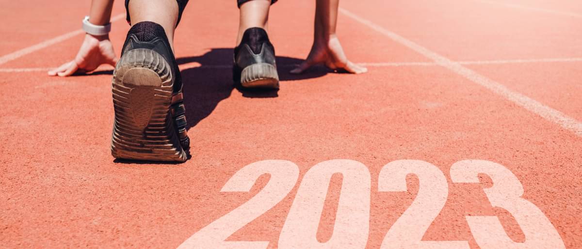 Eine Läuferin kniet an der Startlinie auf einer Aschebahn, man sieht sie von hinten und das neue Jahr mit der Zahl "2023" ist davor zu lesen.