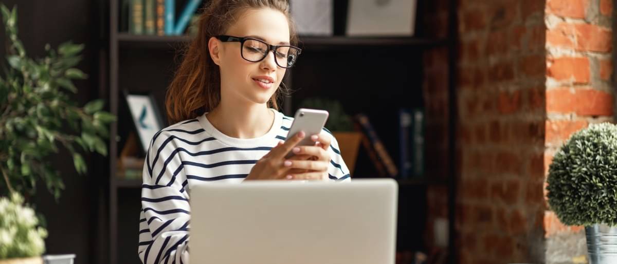 Eine junge Frau sitzt am Schreibtisch und macht Online Banking. Zur Freigabe verwendet sie das photoTAN Verfahren und scannt den Code auf dem Laptop mit ihrem Smartphone ab.