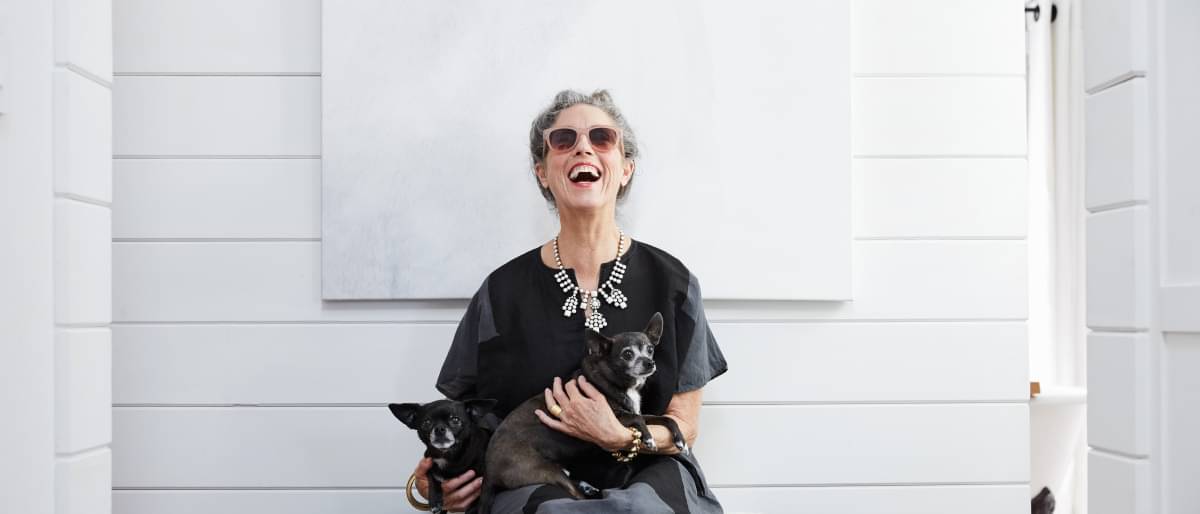 Altere Frau, die auf einer Bank sitzt und mit ihren beiden Hunden auf dem Schoß euphorisch lacht