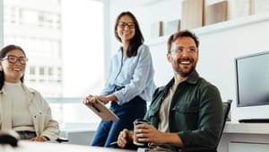 Zwei Frauen und ein Mann sitzen in einem Büro am Tisch. Alle freuen sich und blicken lächelnd in die selbe Richtung.