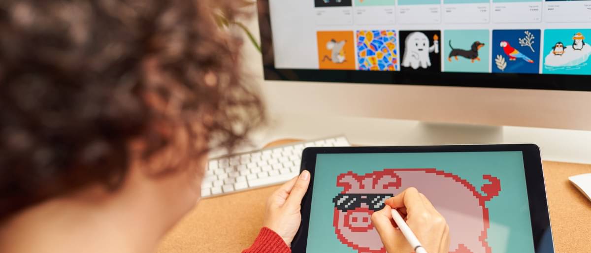 Eine Künstlerin zeichnet auf einem Tablet ein rosa Schweinchen mit Sonnenbrille. Im Hintergrund sind weitere Werke von ihr auf einem Monitor zu sehen.