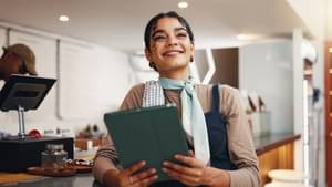 Eine Frau lehnt in einer Bar am Tresen und hält ein Tablet in den Händen. Sie lächelt und blickt in die Ferne.