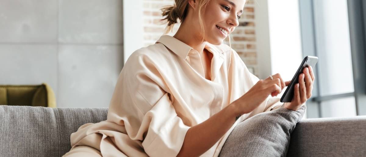 Eine blonde junge Frau in heller Kleidung sitzt gemütlich auf dem Sofa und liest auf ihrem Smartphone.