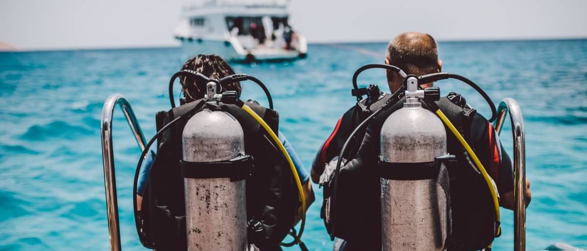 Zwei Taucher mit Sauerstoffflaschen und Tauchanzügen sitzen am Ende eines Bootes und machen sich bereit ins Meer zu gleiten, um zu tauchen.