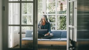 Frau mit grauen langen Haaren sitzt im Schneidersitz auf dem Sofa am Fenster und arbeitet am Laptop in ihrem Schoß.