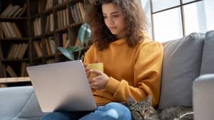 Junge Frau mit lockigen Haaren sitzt mit ihrer Katze auf einem Sofa. Sie hat ihren Laptop auf dem Schoß und hält eine Kaffeetasse in der Hand