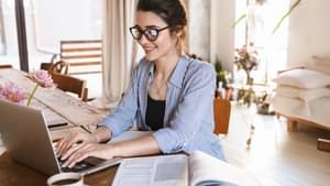 Junge Frau mit Dutt und Brille sitzt an ihrem Schreibtisch, auf dem sich einige Unterlagen stapeln, während sie an ihrem Laptop arbeitet.
