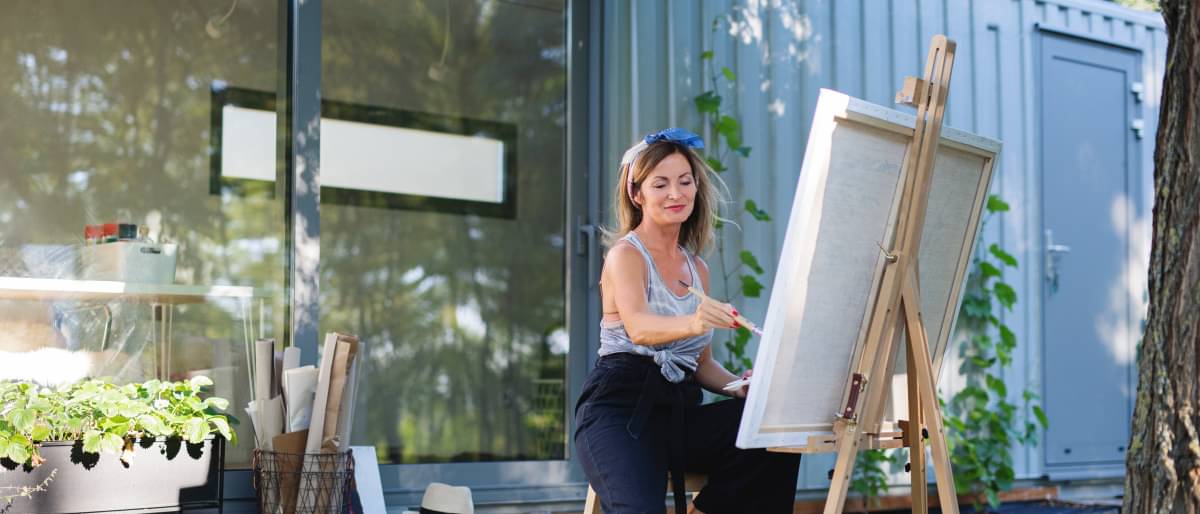 Ältere Frau sitzt lässig auf einem Hocker vor ihrem Atelier und malt an einer Staffelei.