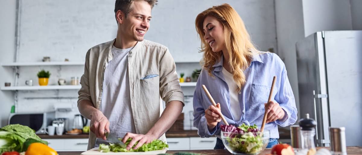 Ein Paar steht in einer großen, hellen Küche und bereitet gemeinsam ein gesundes Gericht zu. Der Mann schneidet Salatblätter, während die Frau Salat in einer Schüssel umrührt.