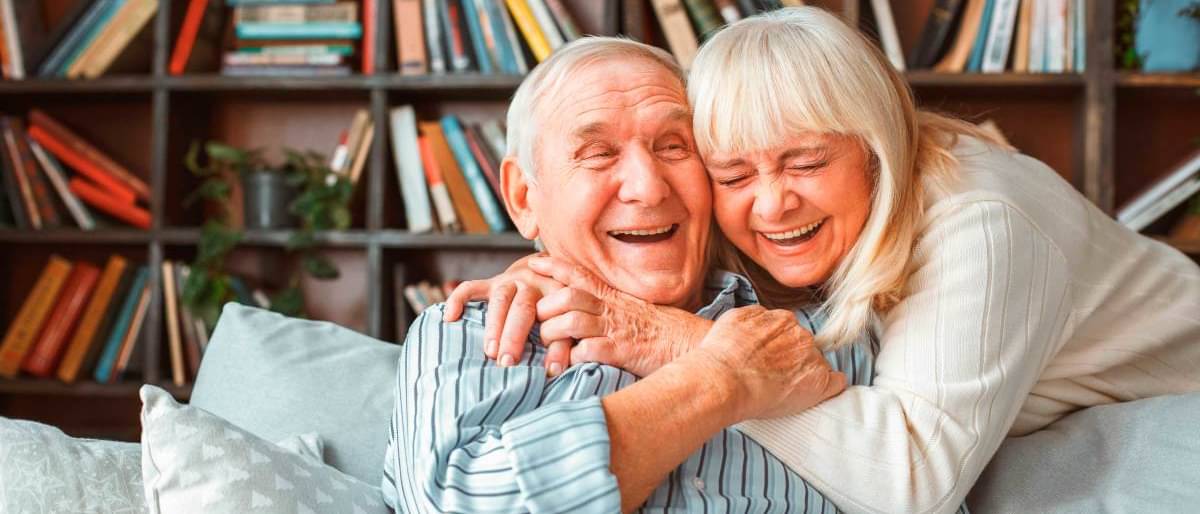 Ein älteres Pärchen im Wohnzimmer umarmt sich lachend - Altersgerechtes Wohnen