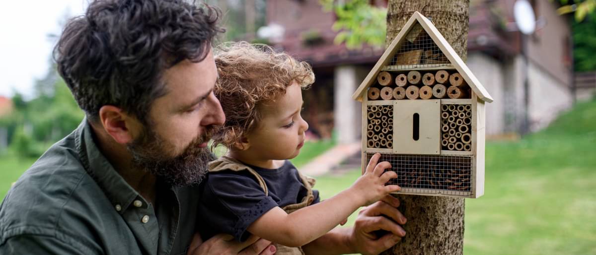 Ein Vater hebt seine kleine Tochter hoch, um ihr ein Insektenhotel zu zeigen.