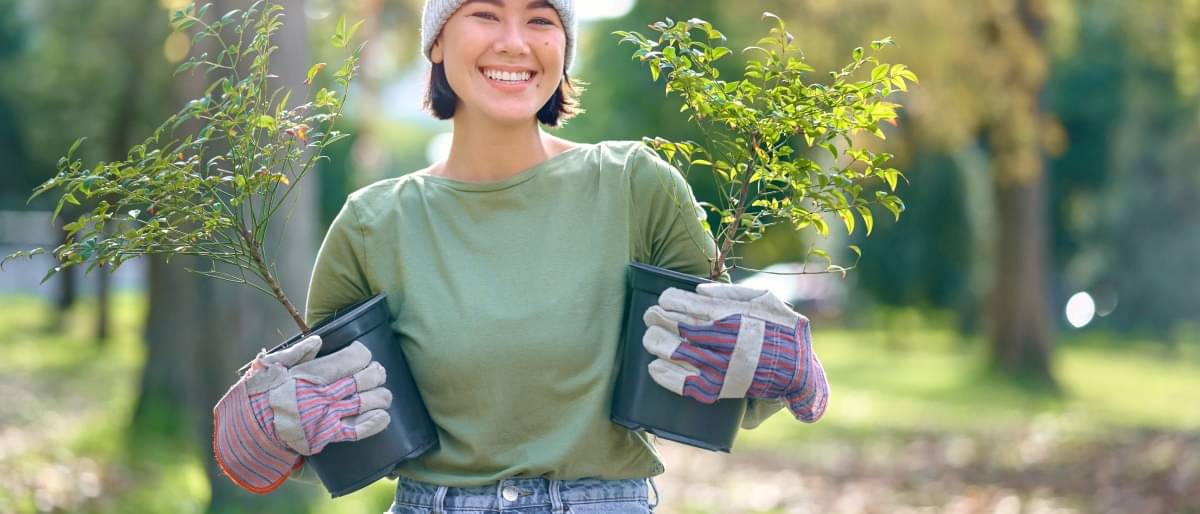 A woman wearing a hat and gardening gloves stands in a park and holds two small tree saplings in her arms. She smiles at the camera.
