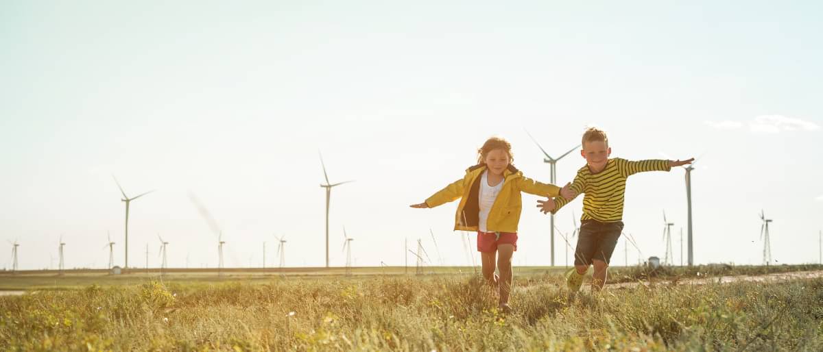 Zwei Kinder laufen über grüne Wiese. Im Hintergrund erkennt man Windkraftanlagen. Ein guter CO2 Fußabdruck