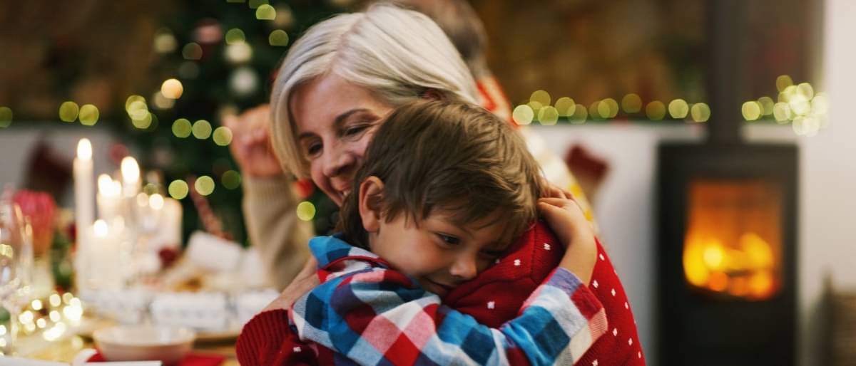 Ein Kind umarmt eine Frau vor einem Weihnachtsbaum und schaut glücklich und zufrieden
