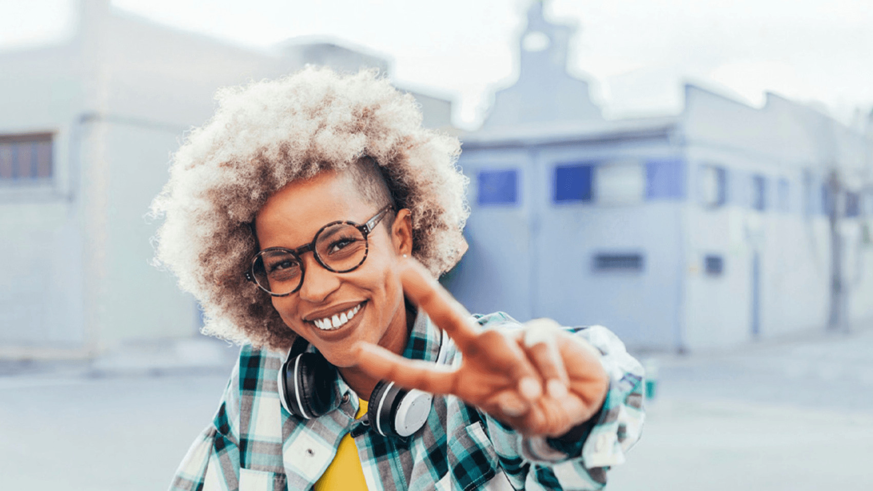 Frau mit Brille und Kopfhörern um den Hals. Sie streckt ihre Hand zu einem Peace-Zeichen aus und lächelt dabei freundlich