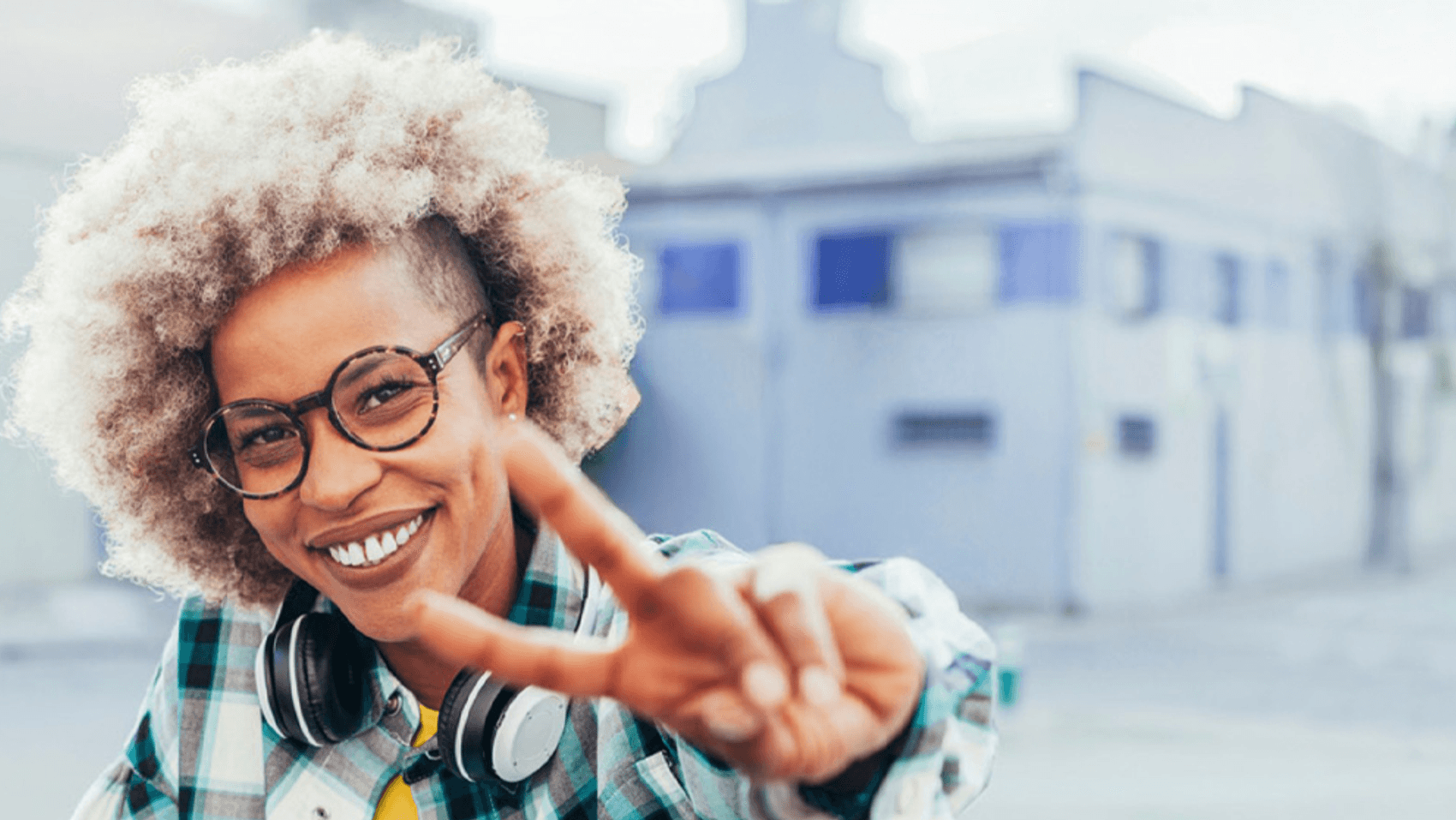 Frau mit Brille und Kopfhörern um den Hals. Sie streckt ihre Hand zu einem Peace-Zeichen aus und lächelt dabei freundlich