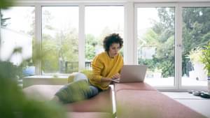 Eine junge, dunkelhaarige Frau sitz auf einem Sofa, arbeitet entspannt am Laptop.