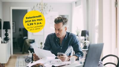 Junger Mann sitzt an Tisch vor Laptop und hat Unterlagen und Handy in den Händen: Bis 5,99 % p.a.